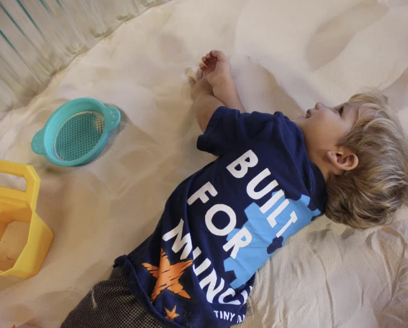 A child in a Built for Muncie Tiny Trades shirt lying back in the sand, smiling