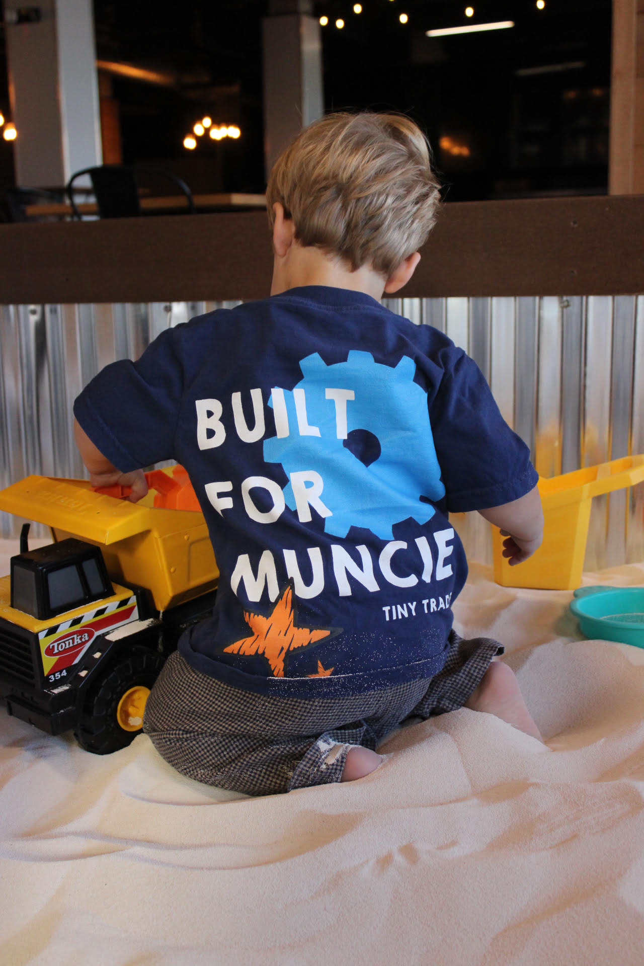 A child in a Built for Muncie Tiny Trades shirt playing with a toy dump truck in the sandbox